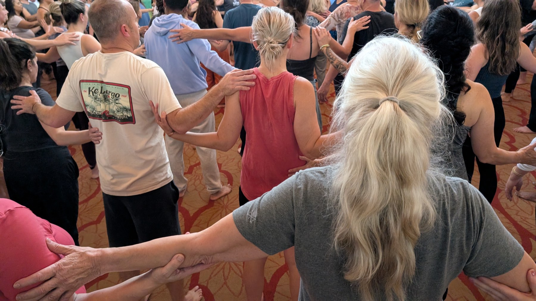 A room of people standing in a chain, each with a hand resting on the shoulder or arm of the person in front of them.