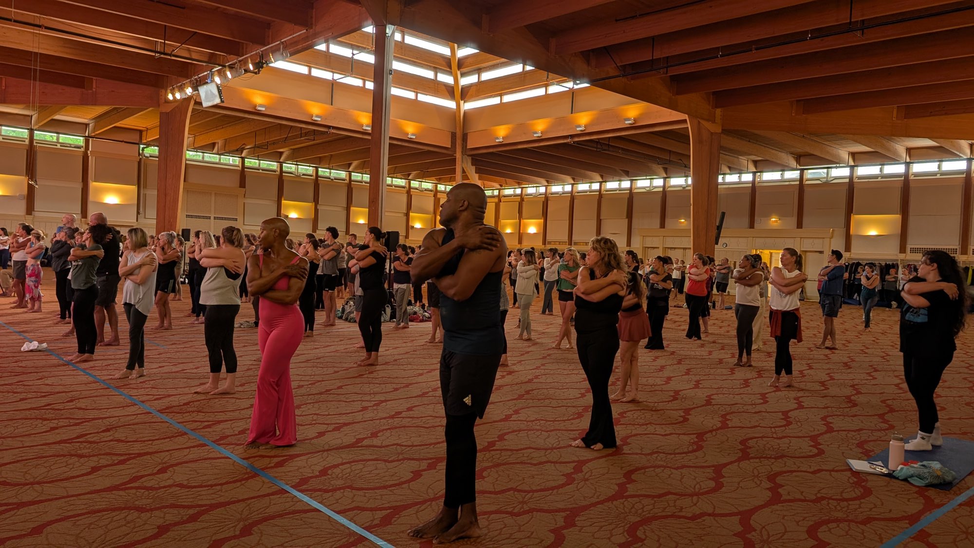 Hundreds of people spread across a large timber-framed hall, each holding the same self-applied shoulder maneuver.