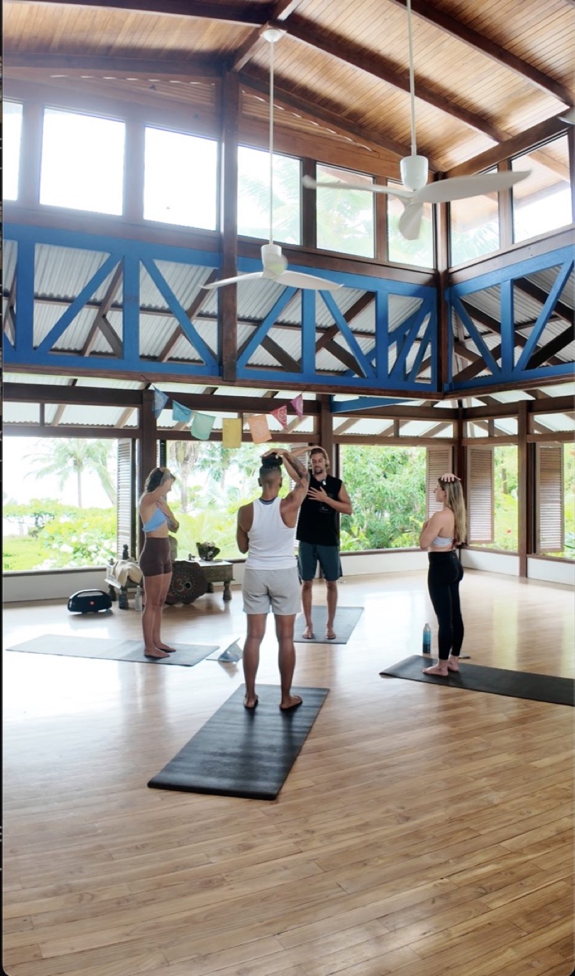 Four people standing on mats in an open-air timber pavilion, mirroring a coach who holds one hand to his head and one to his heart.